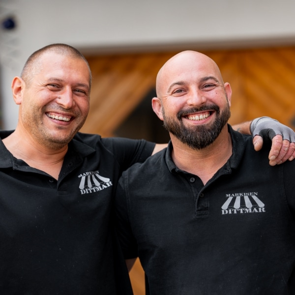 Zwei lächelnde Männer mit rasierten Köpfen und schwarzen Hemden stehen im Freien, haben die Arme um die Schultern des anderen gelegt und verkörpern die für eine Fachbetrieb-Montage typische Kameradschaft.