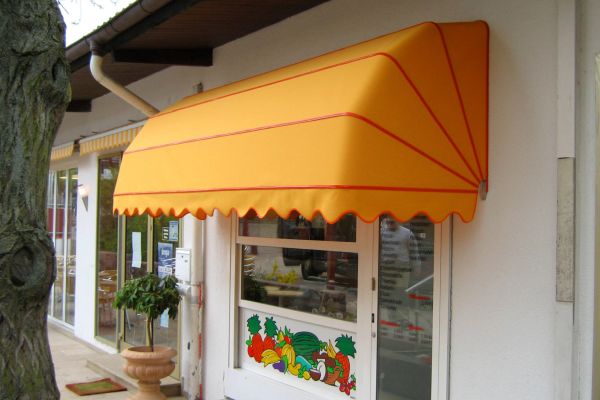 Ladenfront mit einer gelben, gewellten Markise, die an traditionelle Markisendesigns erinnert, über einer mit Obst- und Gemüseillustrationen verzierten Tür.