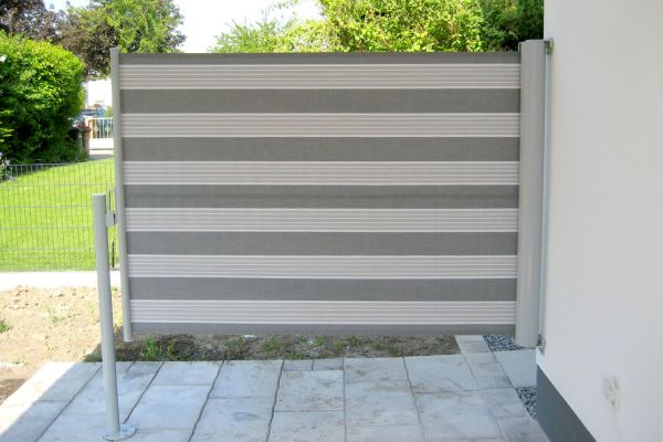 Ein grau-weiß gestreifter, ausziehbarer Sichtschutz aus der Fertigung eines Fachbetriebs ziert die Terrasse mit Eleganz, während üppiges Grün einen natürlichen Hintergrund bildet.