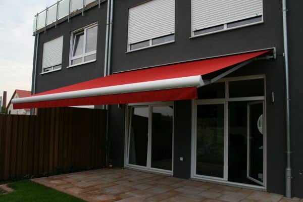 Eine rot-weiße, einziehbare Markise erstreckt sich elegant über die Terrasse und ergänzt die moderne Fassade und die großen Fenster eines zeitgenössischen Gebäudes.