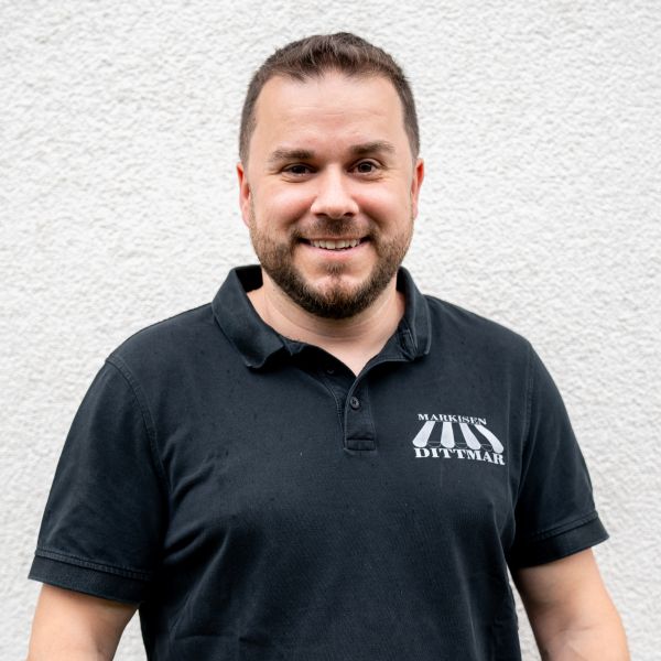 Ein lächelnder Mann in einem schwarzen Poloshirt mit aufgedrucktem Text steht vor einer strukturierten weißen Wand und strahlt das Selbstvertrauen von jemandem aus, der gerade die perfekte Marke für eine optimale Beschattung gefunden hat.