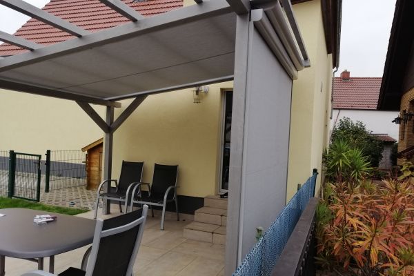 Terrasse mit Pergola bietet fachmännische Beschattung, Stühle und Tisch auf gefliestem Boden. Fachbetrieb-Einbau führt zu einer Haustür, umgeben von Büschen und bietet Ausblick auf das Nachbarhaus.