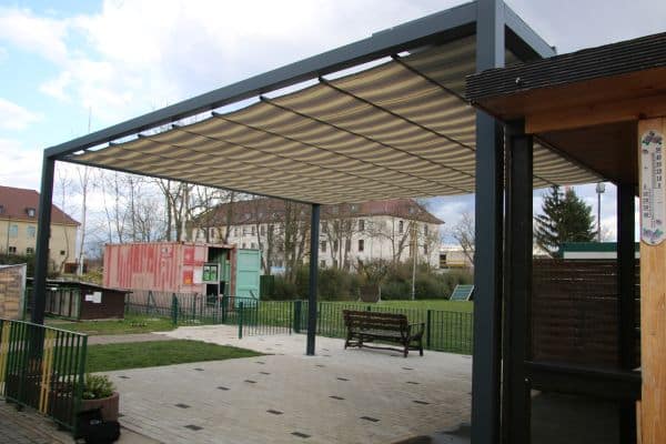 Eine Metallpergola mit Stoffdach sorgt für die Beschattung eines Terrassenbereichs, ergänzt durch eine Sitzbank und üppige Grünflächen in einem Wohnumfeld.
