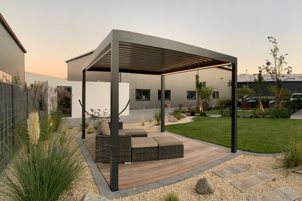 Eine moderne Außenpergola mit bequemen Sitzgelegenheiten ruht auf einem Holzdeck, aufgewertet durch die Beschattung einer eleganten Markise. Die Kulisse wird bei Sonnenuntergang durch einen angelegten Garten wunderschön ergänzt.