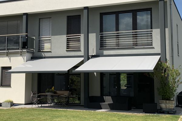 Eine moderne Hausfassade verfügt über zwei Balkone und ausfahrbare Markisen für eine perfekte Beschattung, gepaart mit Terrassenmöbeln auf einer gepflegten Rasenfläche.