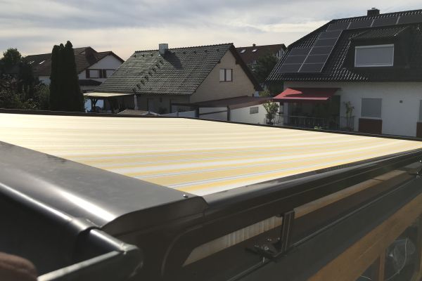 Eine gelb-weiß gestreifte Markise erstreckt sich über das Dach und bietet eine hervorragende Beschattung vor dem bewölkten Himmel, mit charmanten Häusern im Hintergrund.