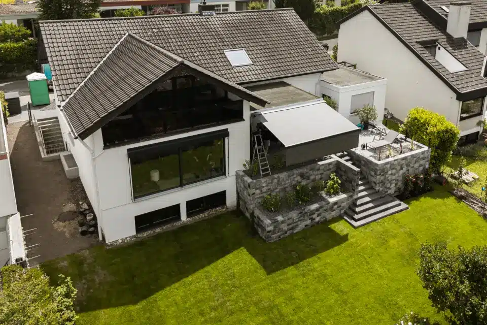 Luftaufnahme eines modernen Hauses mit Schrägdach und großen Fenstern, ergänzt durch eine abgestufte Gartenterrasse mit Sitzbereich. Fachmännische Beschattungslösungen sorgen für die perfekte Balance aus Licht und Komfort, fachgerecht installiert von einem vertrauenswürdigen Fachbetrieb.