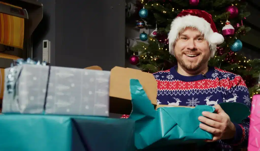 Mann mit Weihnachtsmütze und festlichem Pullover, wie in einer Szene aus einer fröhlichen Montage, öffnet eifrig Geschenke neben dem Weihnachtsbaum.