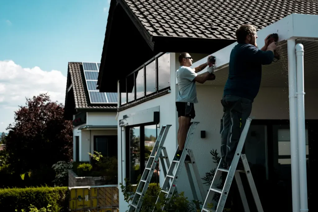 Zwei Männer auf Leitern reparieren fachmännisch die Dachtraufe eines modernen Hauses, das mit Solarmodulen auf dem Dach und einer eleganten Markise zur Beschattung ausgestattet ist, und das alles unter einem klaren Himmel.
