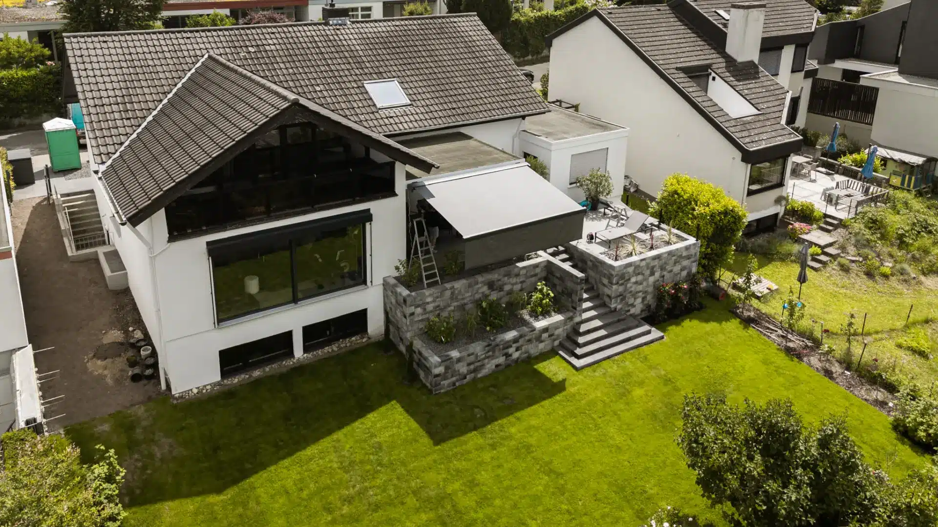 Luftaufnahme eines modernen Hauses mit angelegtem Garten, Steinterrasse und großen Fenstern, die für eine optimale Beschattung fachmännisch entworfen wurden.