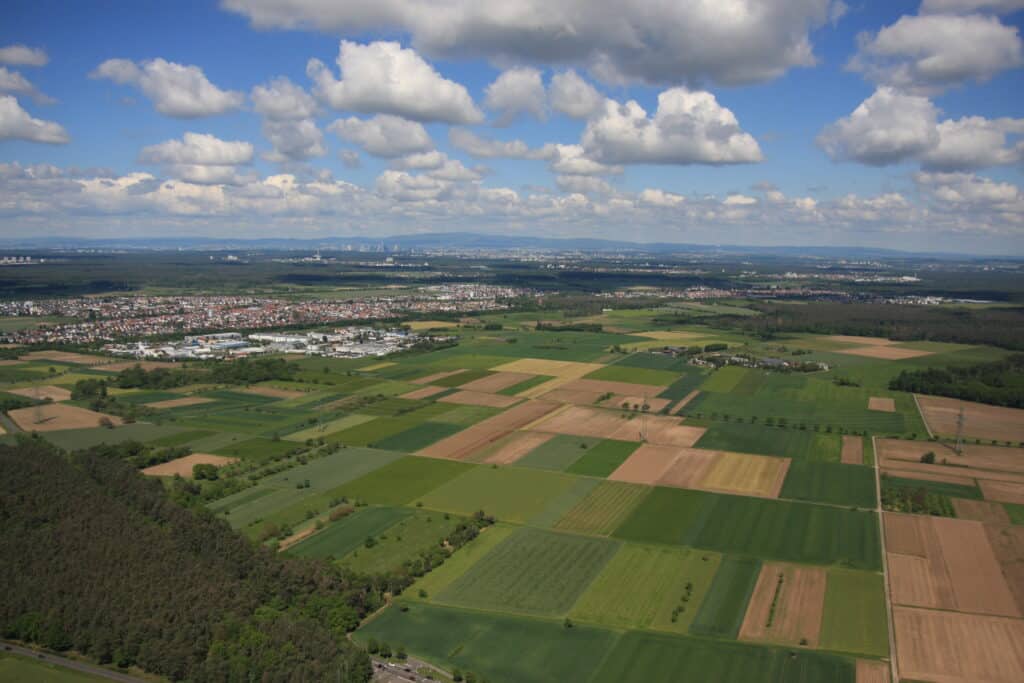 Luftaufnahme von Ackerland, Feldern und einer entfernten Stadt unter einem teilweise bewölkten Himmel – die perfekte Kulisse für alle, die in Römerberg Markise kaufen möchten.