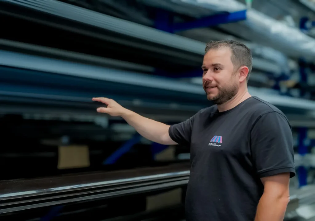 Ein Mann in einem schwarzen Hemd gestikuliert in Richtung gestapelter Metallträger in einem Industrielager.