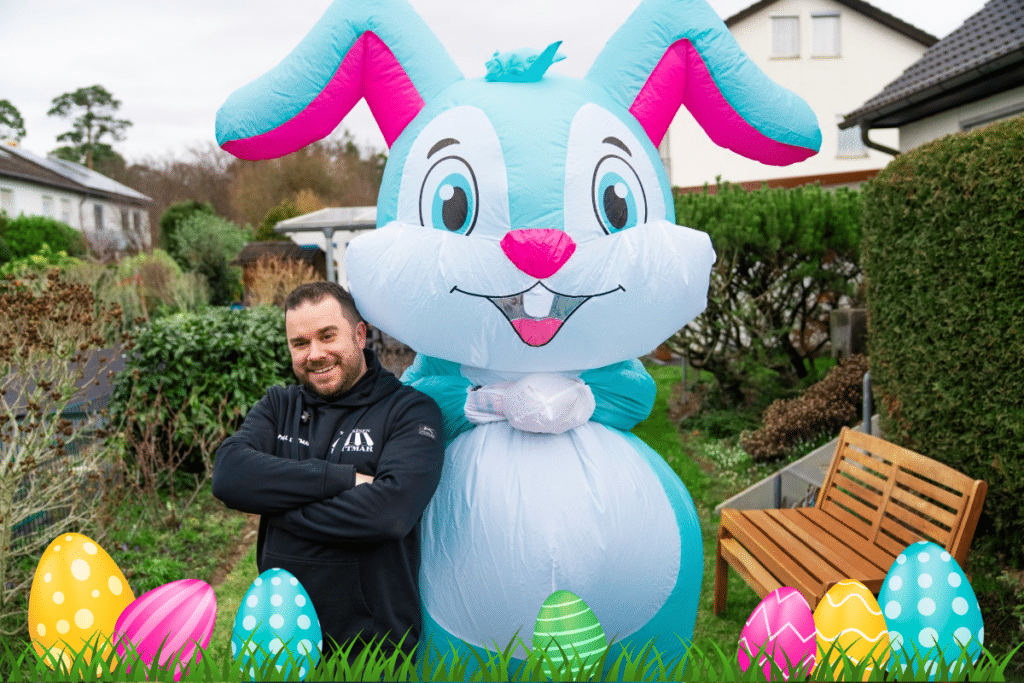 Ein Mann steht lächelnd neben einem großen aufblasbaren Hasen, vor dem bunte Ostereier im Gras liegen.